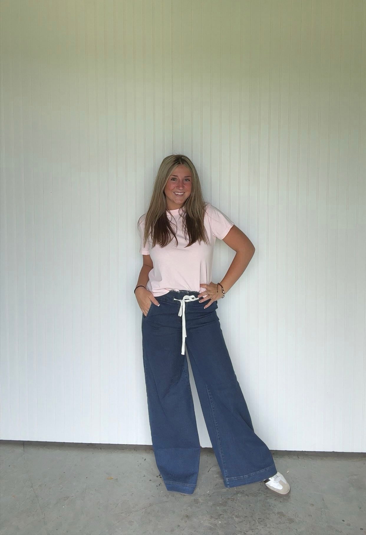 Person wearing a pink shirt and blue jeans standing against a white wall.