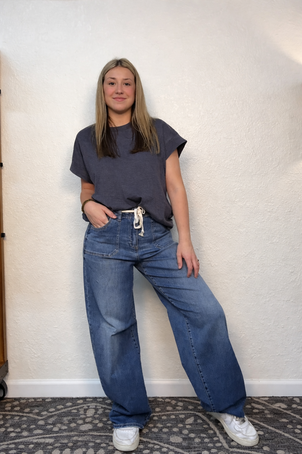 Person wearing a dark gray t-shirt and blue jeans standing against a white wall.