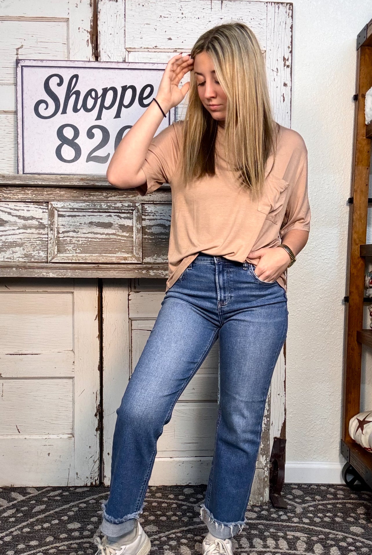 Woman wearing a beige shirt and blue jeans standing in a room with a rustic wooden shelf and decorative sign.