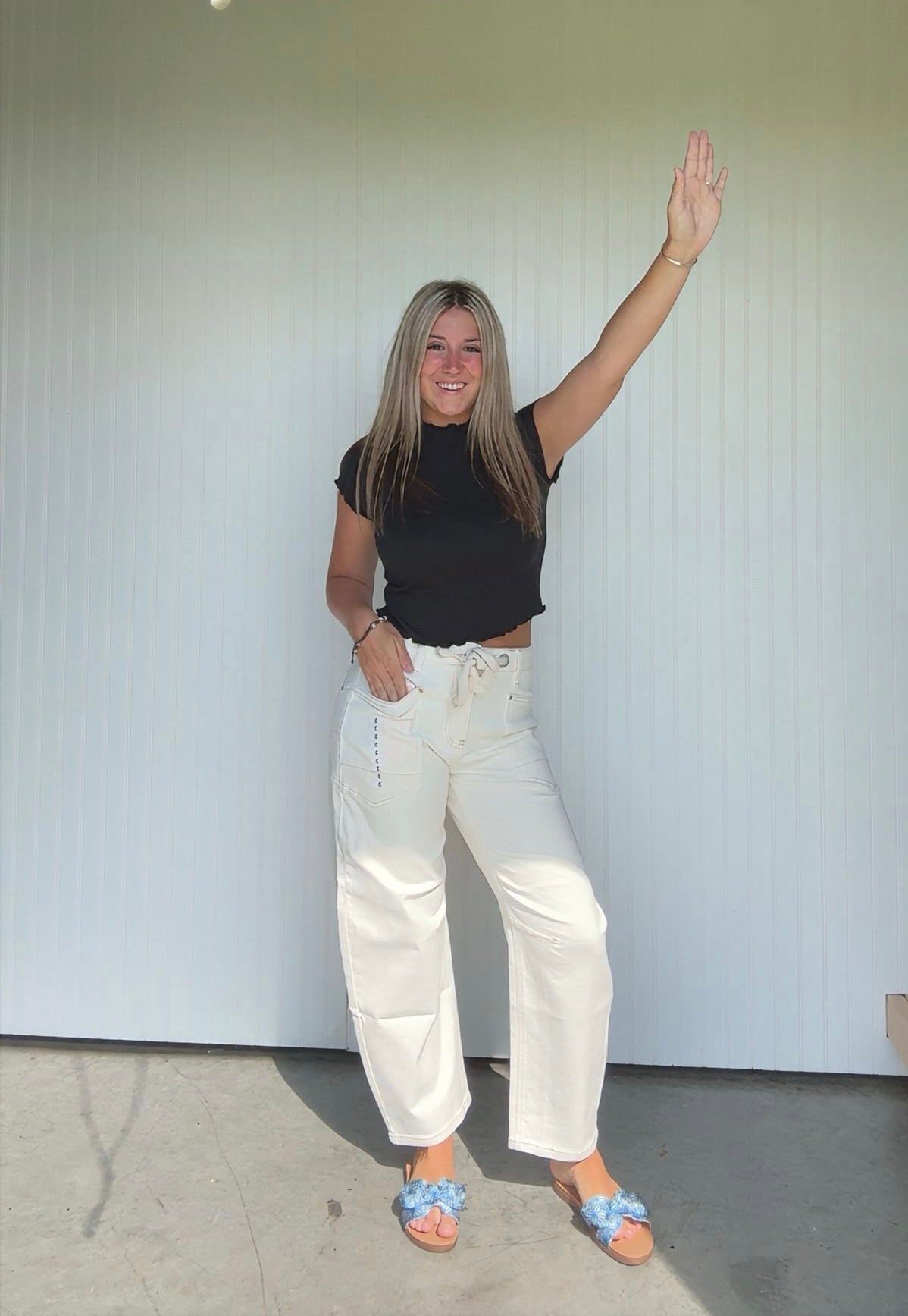 Woman wearing a black top and white pants waving in front of a white wall.