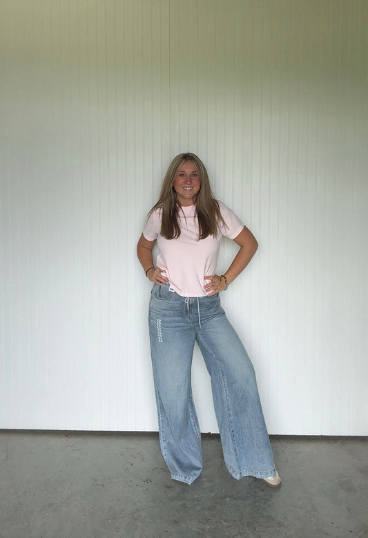 Person wearing a pink shirt and blue jeans standing against a white wall.